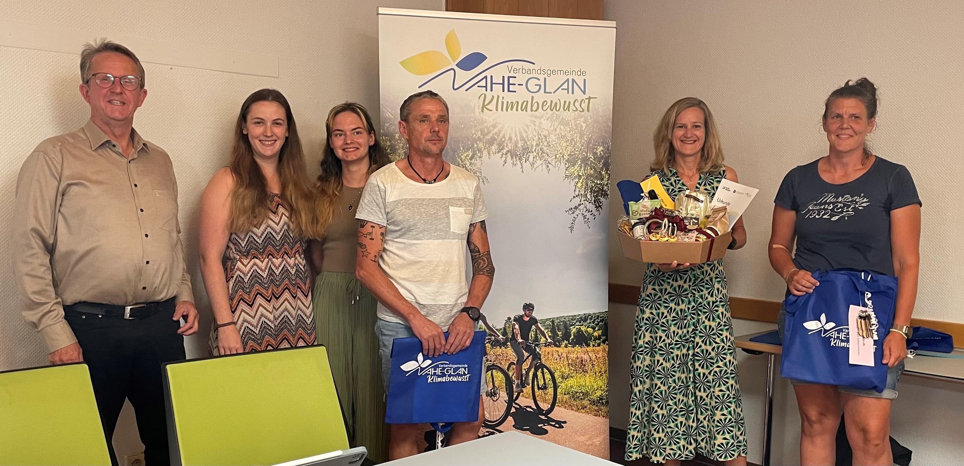Sechs Personen mit Präsenten in den Händen vor einem Banner mit der Aufschrift Verbandsgmeinde Nahe-Glan klimabewusst und Radfahrern