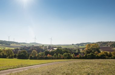 Foto aus entfernter Perspektive mit Blick über die Hausdächer der Ortsgemeinde Bärweiler, grüne Vegetation, blauer wolkenloser Himmel bei hochstehender Sonne, am Horizont einige Windräder.