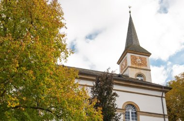 Foto eines eher schlichten Kirchengebäudes umrahmt von grün belaubten Bäumen bei heiter bewölktem Himmel.