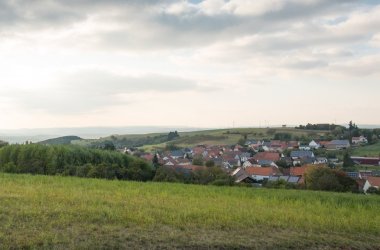 Foto aus Blickrichtung eines begrünten Hügels auf eine im leichten Tal gelegene Siedlung. in der oberen Bildhälfte ein leicht bewölkter Himmel mit von links scheinender, tiefstehender Sonne.