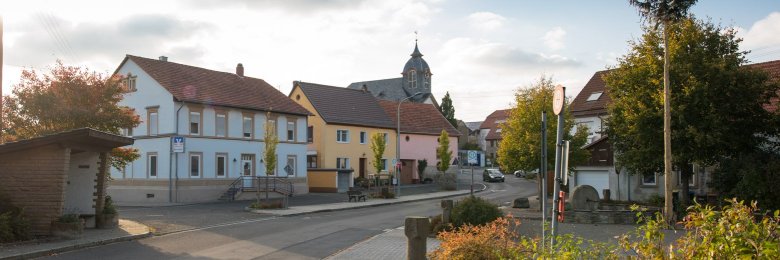 Foto einer Straßenkreuzung innerhalb einer kleinen Siedlung. Fünf Häuser und im Hintergrund ein Kirchengebäude erkennbar. Im Bildmittelpunkt eine Bushaltestelle vor drei Häusern unterschiedlich gefärbter Fassaden. Vereinzelt grün belaubte Vegetation.