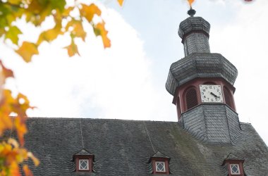 Foto eines Daches und Turmes eines Kirchengeäudes vor leicht bewölktem Himmel. Im Vordergrund der linken Bildhälfte grünes Laub eines Baumes.