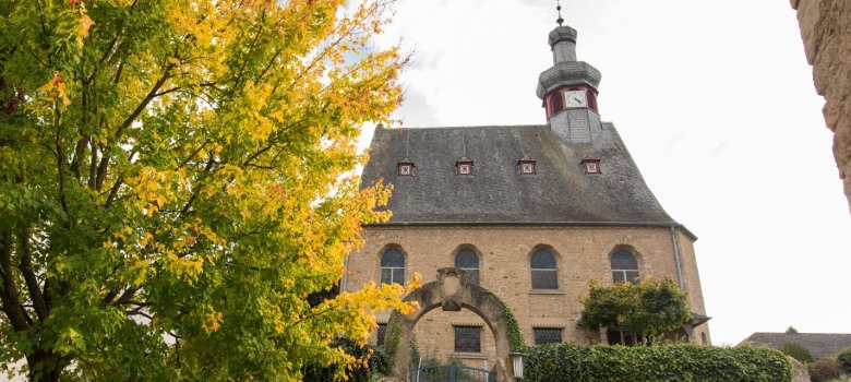 Foto einer Kapelle hinter einer hohen Hecke mit steinernem Torbogen. Im Vordergrund in der linken Bildhälfte ein grün belaubter Baum.