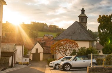 Foto einer Kapelle an einer nach hinten abfallenden, schmalen Straße mit wenigen Häusern und drei Autos entgegen einer teifstehenden, am Horizont verschwindenden Sonne bei leicht bewölktem Himmel.