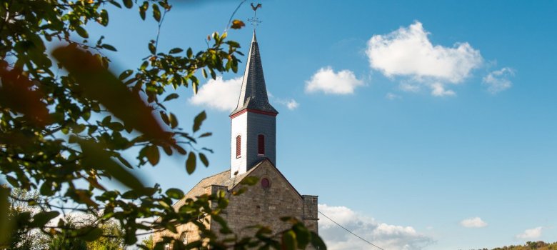 Foto des oberen Teils eines Kirchengebäudes hinter einem grün belaubten Baum, vor kaum bewölktem Himmel.