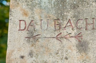 Foto Nahaufnahme eines Steins mit der eingemeisselten Inschrift "Daubach" und einem nach links gerichteten Pfeil darunter.