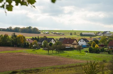 Foto einer kleinen Siedlung etwas vertieft, naturumschlossen in einer grünen leicht hügeligen Feld- und Wiesenlandschaft mit nur wenigen Bäumen und Sträuchern liegend bei leicht bewölktem Himmel.