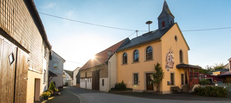 Foto eines Straßenzuges mit beidseitigen Häuserreihen und einem kleinen Kirchengebäude bei wolkenlosem Himmel und strahlender Sonne.