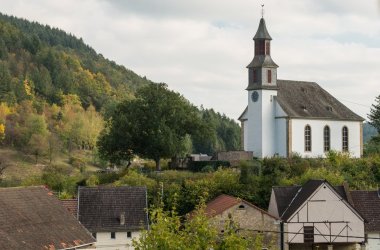 Foto eines Kirchengebäudes hinter und über einer kleinen Siedlung neben einem grün bewladetem Hügel.