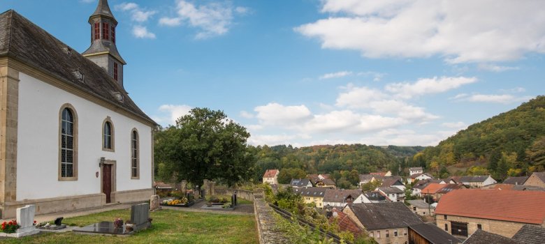 Foto eines kleinen Friedhofs mit sechs Grabstellenneben einem Kirchengebäude. Im Hintergrund Gebäude und Gebäudedächer einer kleinen Siedlung.