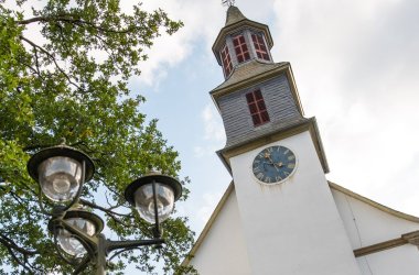 Foto aus Froschperspektive eines Kirchengebäudes hinter einen dreiarmigen Straßenlaterne und einem grün belaubten Baum vor bewölktem Himmel.
