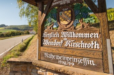 Foto eines Holzschildes mit dem Wappen und Namen der Ortsgemeinde Kirschroth und der Naheweinprinzessin von 1998 zwischen einem weinbewachsenem Hügel und der Zufahrtsstrße zu einer Siedlung.