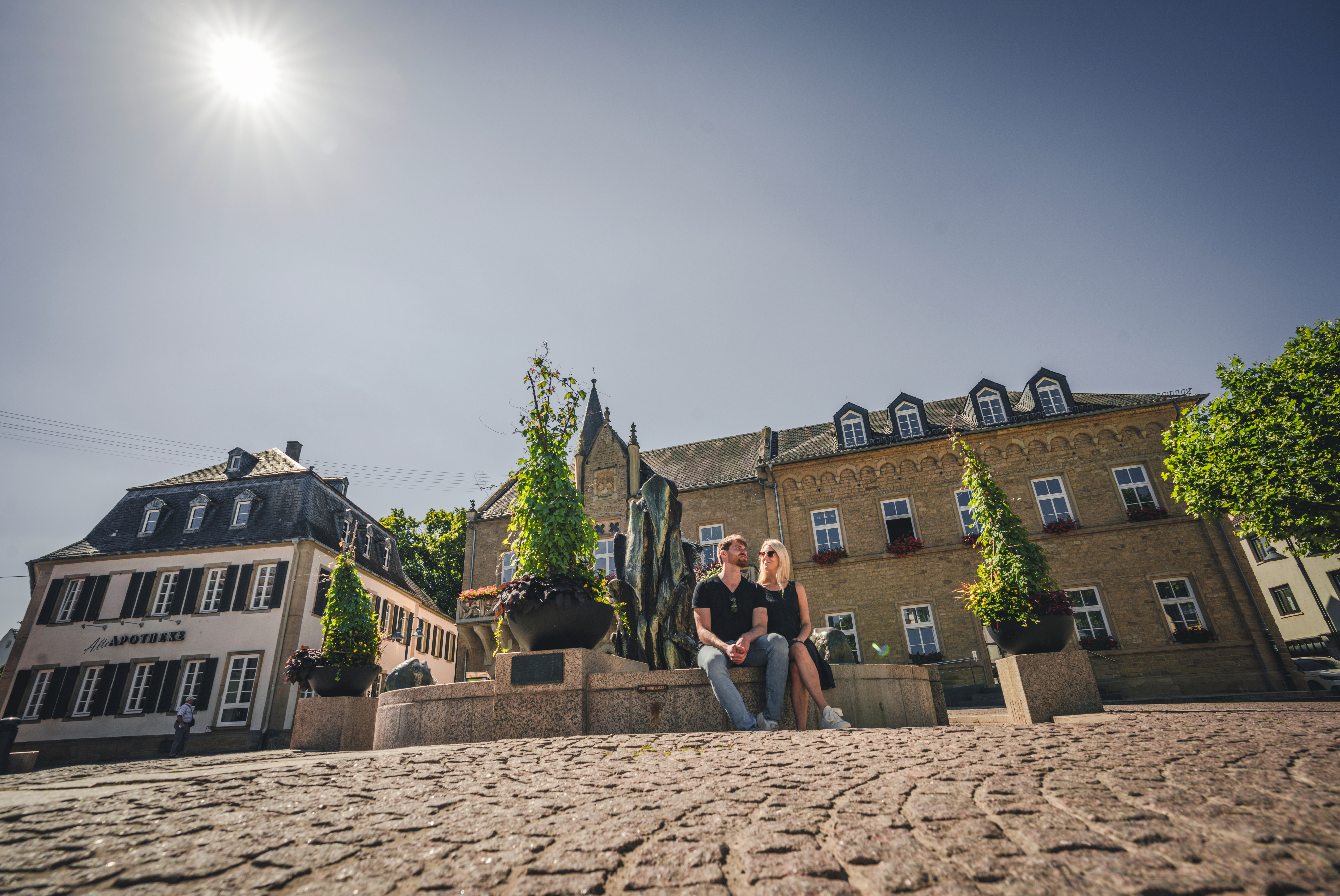 Marktplatz mit Brunnen auf dessen Rand ein Mann und eine Frau in der Sonne sitzten