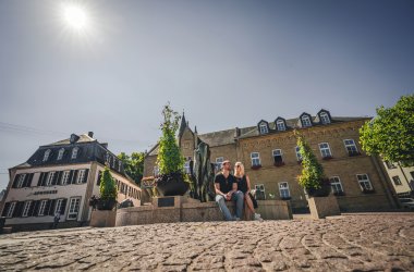 Marktplatz mit Brunnen auf dessen Rand ein Mann und eine Frau in der Sonne sitzten