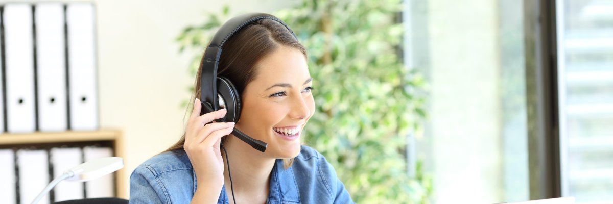 Symbolbild einer Service-Mitarbeiterin zeigt eine freundlich lächelnde Frau mit Headset vor einem Aktenregal sitzend an einem Schreibtisch mit offenem Laptop darauf.
