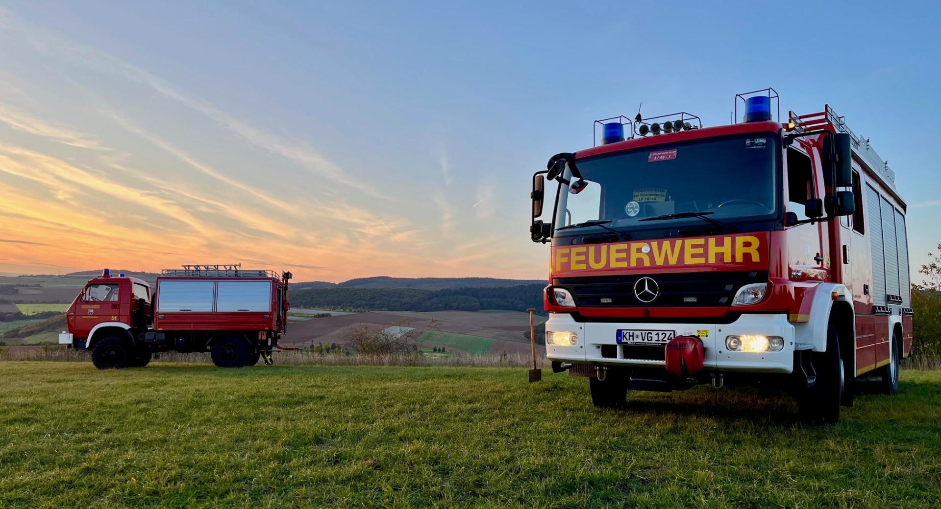 Zwei Feuerwehrfahrzeuge auf einer grünen Wiese vor Weinbergen im Sonnenuntergang.