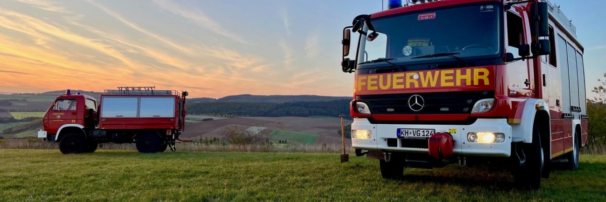 Zwei Feuerwehrfahrzeuge auf einer grünen Wiese vor Weinbergen im Sonnenuntergang.