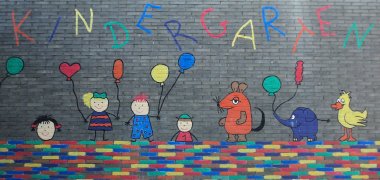 Eine bunt bemalte Steinmauer mit der Aufschrift Kindergarten, skizzenhaft dargestellter Kinder, der Maus, dem Elefanten und der Ente aus der Sendung mit der Maus.