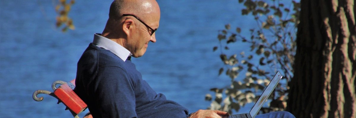 Nahaufnahme einer auf einer Bank vor einem Gewässer unter einem Baum sitzenden Person mit Blick auf einen offen auf dem Schoß liegenden Laptop.