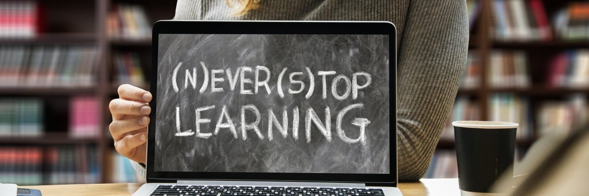 Symbolbild. Ein aufgeklappter Laptop auf einem Tisch mit einem Pappbecher vor einer Person vor einem Bücherregal zeigt Tafelkreidenaufschrift "Never Stop Learning", wobei der Buchstabe "N" zu Beginn des Wortes "Never" und der Buchstabe "S" zu Beginn des Wortes "Stop" in Klammern stehen, um eine Doppeldeutigkeit darzustellen.